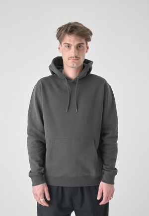 Junger Mann mit kurzen braunen Haaren und Schnurrbart, trägt einen dunkelgrauen Hoodie und schwarze Hose, steht vor einem schlichten hellen Hintergrund.