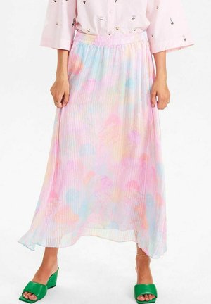 Femme portant une jupe midi plissée de couleur pastel avec des motifs abstraits, un chemisier rose pâle et des sandales compensées vertes à bout ouvert.