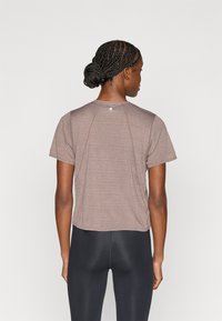 Kortermet, brun treningsskjorte i teksturert materiale med rund hals og subtil logo på baksiden. Kombinert med svarte leggings.