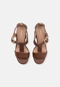 Sandalias de cuero marrón con suela plana, que presentan correas anchas, detalles de hebilla dorada y un diseño de espalda abierta para ponerlas y quitarlas con facilidad.