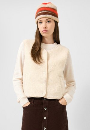 Jonge vrouw die een gestreepte wollen muts draagt, een crèmekleurig fleecevest, een lichte trui en een bruine corduroy rok met knopen, met handen in de zakken.