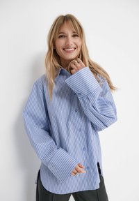 Niebiesko-biała koszula w paski z długimi rękawami, dużymi mankietami, guzikami z przodu i rozcięciami po bokach. Materiał wydaje się mieć teksturę.