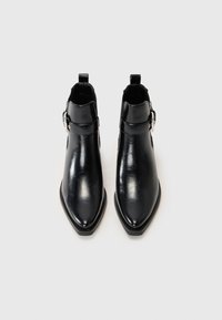 Botines negros con punta afilada, hechos de cuero brillante. Presentan una hebilla lateral y una textura suave con un diseño minimalista.