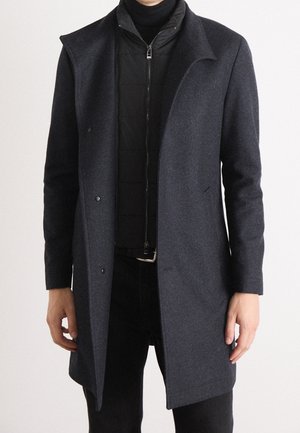 Mężczyzna ubrany w ciemnoszary płaszcz nałożony na czarną kurtkę na zamek i czarny golf, stoi na tle jednolitego białego tła.