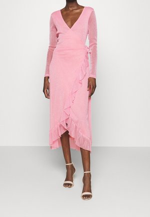 Femme portant une robe portefeuille rose scintillante à manches longues avec un ourlet asymétrique à volants, associée à des talons nude à bride cheville.