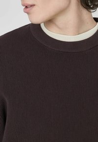 Sweater côtelé marron foncé avec un col rond, superposé à un t-shirt de couleur claire. Tissu texturé avec des motifs côtelés visibles.
