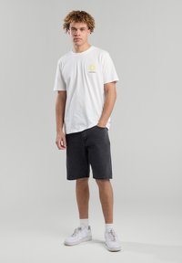 Valkoinen puuvillapaita, jossa on keltainen kukkakuva, yhdistetty mustiin farkkushortseihin ja valkoisiin tennareihin. Yksinkertainen, rento muotoilu.