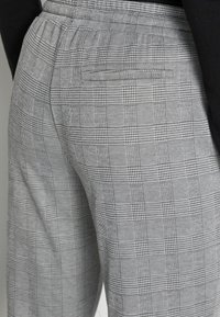 Grijze geblokte broek met een gestructureerd patroon, elastische tailleband en een achterzak, die een uniform rasterontwerp benadrukt.