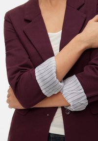 Weinroter Blazer mit Reverskragen, an dessen hochgekrempelten Ärmeln hellblau-weiß gestreifte Manschetten hervorschauen. Zwei Knöpfe sind vorne sichtbar.