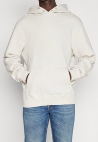 Felpa con cappuccio color crema realizzata in tessuto morbido, con tasca a marsupio frontale, cappuccio regolabile con coulisse e maniche con polsini.