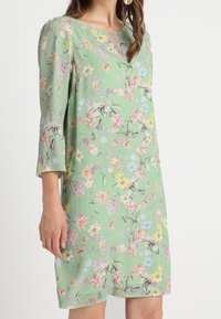 Blommig klänning i ljusgrön tyg, med trekvartsärmar, rund halsringning och en blandning av färgglada blommönster.