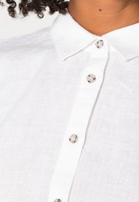 Close-up van een persoon die een wit linnen shirt draagt met een klassieke kraag en vier zichtbare knopen aan de voorkant.