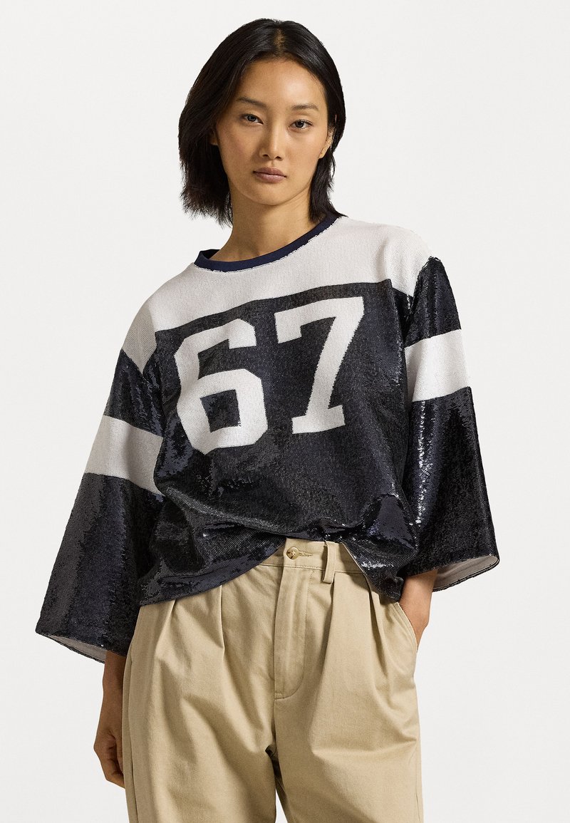 Navy sequined oversized sweatshirt with white stripes and number 67 printed in bold. Paired with beige high-waisted trousers.