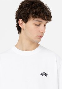 White cotton sweatshirt featuring a round neckline, loose fit, and a small black embroidered logo on the left chest area.