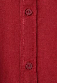 Rotes Hemd mit einer einfachen Leinwandbindung, zwei runden Knöpfen auf der Knopfleiste und sichtbaren Nähten entlang der Kanten.