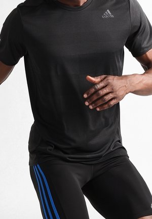 Atleet draagt een zwart Adidas sportshirt en zwarte shorts met blauwe strepen, vastgelegd in een rennende of sprintende pose midden in beweging.