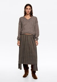 Brązowy sweter z dekoltem w serek o luźnym kroju, zestawiony z długą plisowaną spódnicą w kratę w kolorach brązu, niebieskiego i kremowego, posiadającą boczne rozcięcie oraz brązowe zamszowe buty.