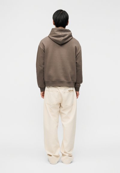 Άτομο που στέκεται με την πλάτη προς τα εμάς, φορώντας καφέ φούτερ με κουκούλα και εκρού παντελόνι φαρδιάς γραμμής με το κέντημα «No Prom» στην πίσω τσέπη.