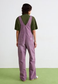 Salopette di cotone lilla con gambe larghe e spalline regolabili, dotata di due tasche posteriori. Indossata sopra una maglietta verde con texture.