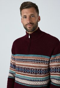 Bordeaux kleurige pullover trui met een hoge hals en een halve rits. Heeft gebande patronen in crème, blauw en bruin over de borst. Wolstructuur.