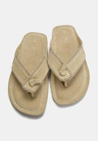 Beige flip-flops i mocka, med ett korslagd remmar med små metallaccenter längs kanterna och en texturerad sula.