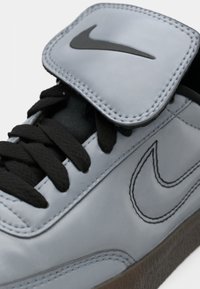 Grå syntetisk sneakers med en svart swoosh-logo, snøring og borrelåsflap øverst. Har svarte lisser og en teksturert såle.