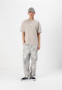 Beige T-Shirt mit Logo auf der Vorderseite, graue gemusterte Hose, schwarze Sneakers. Das Model trägt eine passende graue Mütze und steht vor einem einfarbigen Hintergrund.
