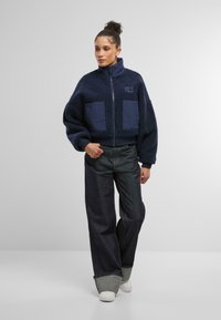 Marinblå fleecejacka med hög krage, dragkedja framtill och två stora tygfickor; kombinerad med lösa mörka denimbyxor och vita skor.