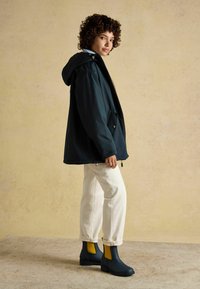 Čierna nepremokavá bunda s kapucňou, nosená cez ľahký sveter a biele nohavice. Oblečenie dopĺňajú námornícke modré a žlté gumové topánky na členky.