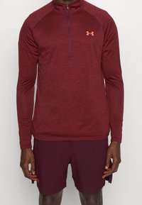 Maroon långärmad atletisk tröja med halvmeterdragkedja och raglanärmar. Har Under Armour-logotyp i orange på bröstet.