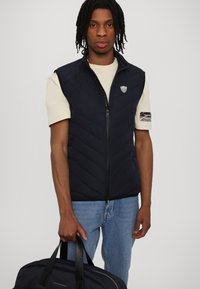 Marineblå quilted vest med glidelås foran og logopatch, brukt over en beige t-skjorte. Modellen holder en matchende marineblå veske med svarte stropper.