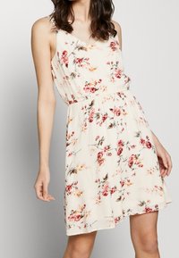 Sukienka kwiatowa w kremowym kolorze z różowymi i zielonymi wzorami. Bez rękawów, dopasowana w talii, z rozkloszowanym dołem i lekką teksturą materiału.