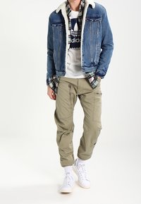 Persona che indossa una giacca di jeans con colletto in pelliccia, camicia a scacchi, t-shirt bianca Adidas, pantaloni cargo color khaki, calzini bianchi e sneakers bianche.