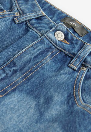 Jean en denim bleu avec des coutures visibles, une fermeture à bouton à l'avant, et une étiquette logo à la taille. Le tissu présente une texture usée.