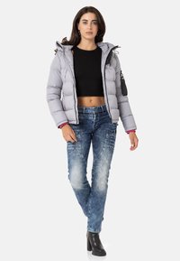 Graue gepolsterte Jacke mit Kapuze, Reißverschluss und schwarzen Akzenten. Kombiniert mit einer ausgewaschenen blauen Jeans und schwarzen Stiefeletten.