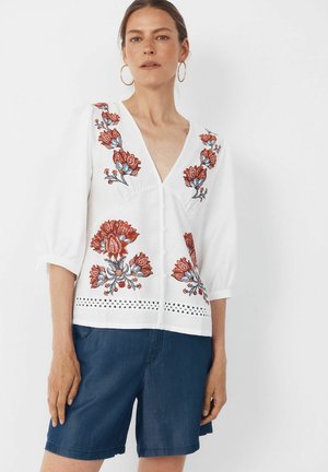 Frau trägt weiße Bluse mit roter und blauer Blumenstickerei und blaue Shorts, steht vor einem schlichten weißen Hintergrund.