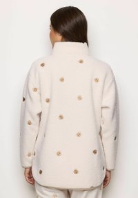 Kvinde iført en hvid fleecejakke med broderede teddy bear-ansigt, vist fra ryggen imod en ensfarvet baggrund.
