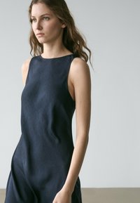 Junge Frau mit langen, welligen Haaren, die ein ärmelloses, dunkelblaues strukturiertes Kleid trägt und vor einem schlichten hellgrauen Hintergrund steht.