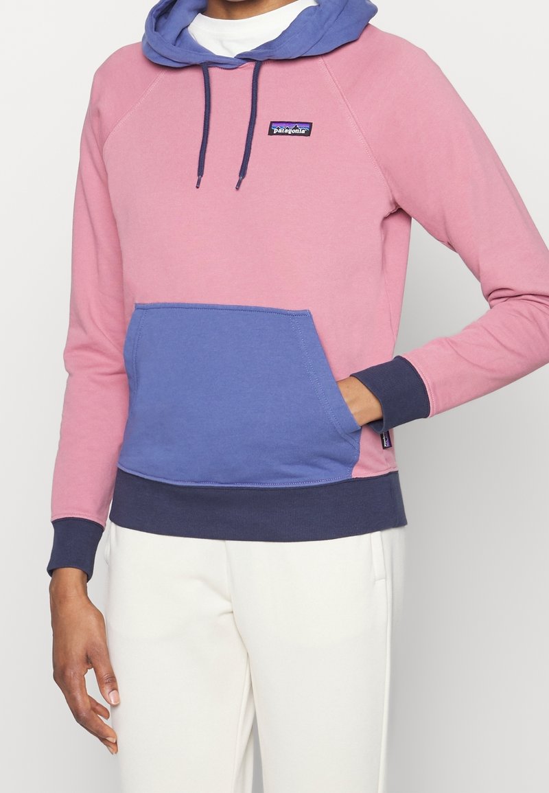 Personne portant un sweat à capuche Patagonia rose et violet avec une poche avant et un pantalon blanc, main détendue sur le côté et dans la poche.
