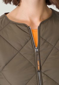 Veste matelassée verte olive avec un col rond, une fermeture éclair argentée et un détail intérieur orange. Étiquette "Think Happy" sur le tirette de la fermeture.