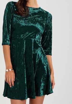 Vrouw draagt een donker groene crushed velvet jurk met driekwart mouwen en een uitlopende rok, geaccentueerd met een parelarmband.
