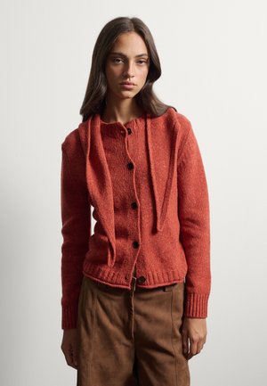 Joven mujer con cabello largo y oscuro que lleva un cárdigan color óxido abotonado y pantalones marrones, de pie frente a un fondo claro uniforme.