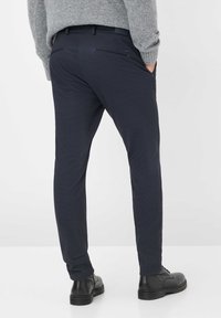 Uomo che indossa pantaloni eleganti blu scuro con tasche laterali e posteriori, stivali di pelle neri e un maglione di lana grigia, mani nelle tasche.