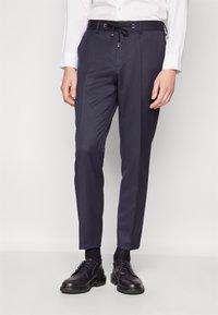 Navy trousers made of fabric with a smooth texture, featuring a drawstring waist, side pockets, and a tailored fit. Worn with black shoes and socks.