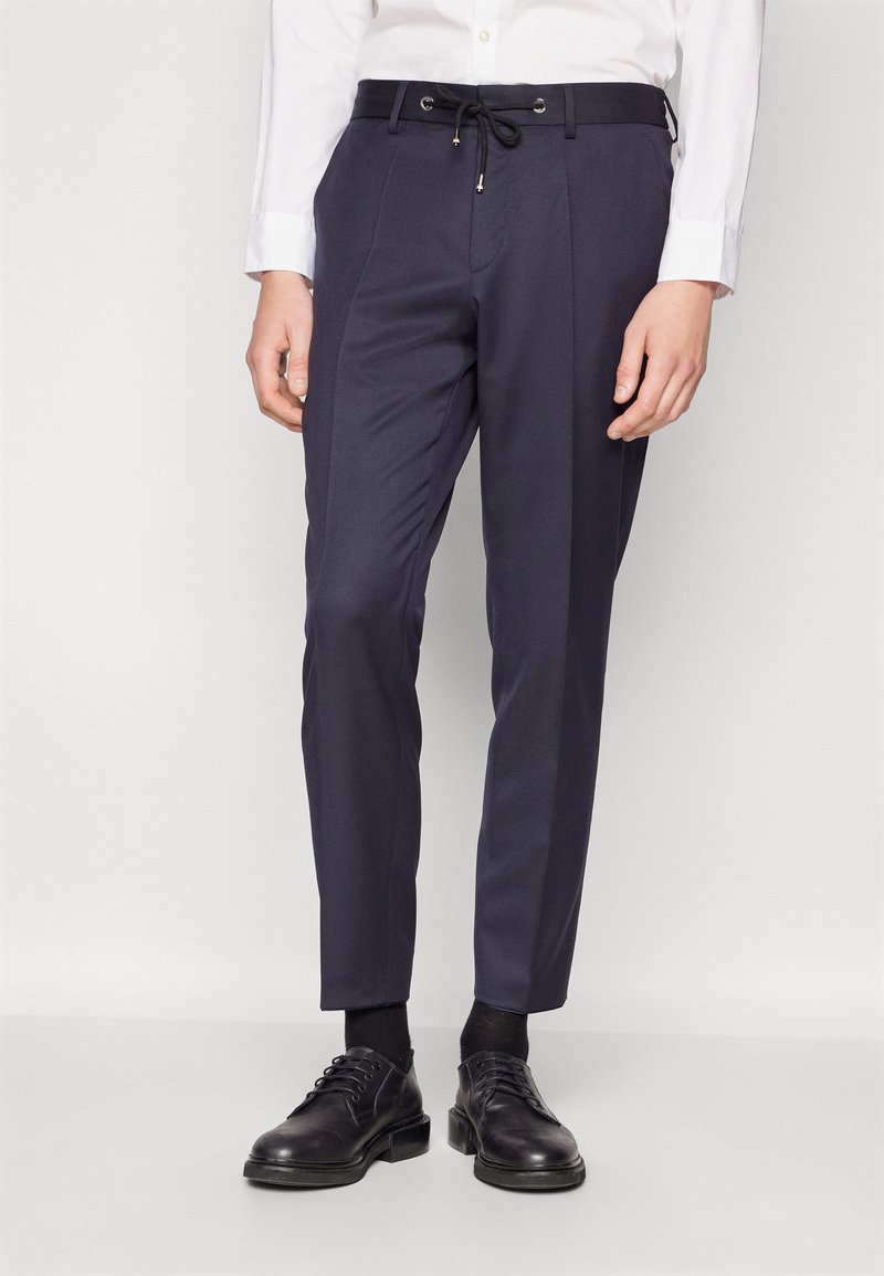Navy trousers made of fabric with a smooth texture, featuring a drawstring waist, side pockets, and a tailored fit. Worn with black shoes and socks.