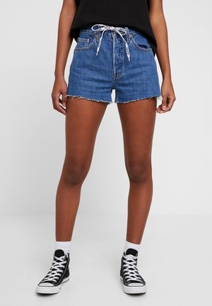 Person trägt blaue Jeansshorts mit ausgefransten Säumen, einen weißen Kordelgürtel, schwarze High-Top Sneaker, weiße Socken und ein schwarzes Top.