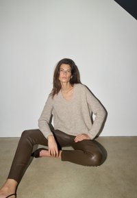 Femme aux longs cheveux bruns assise par terre portant un pull beige et un pantalon en cuir marron foncé devant un mur clair et uni.