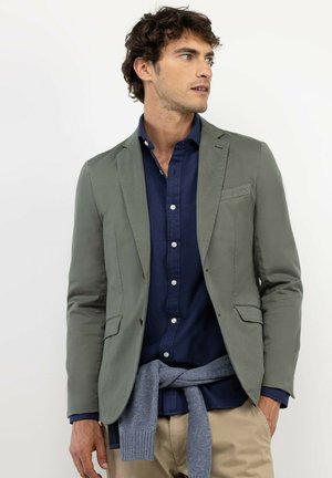Hombre con blazer oliva sobre camisa azul marino y suéter gris atado alrededor de la cintura, mirando hacia un lado sobre un fondo blanco liso.