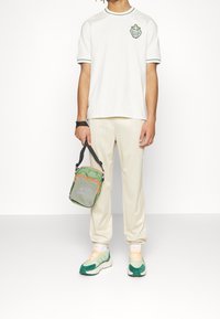 Person som bär en vit t-shirt med liten emblem, beige randiga byxor, gröna och vita sneakers, och håller en grön och orange axelremsväska.