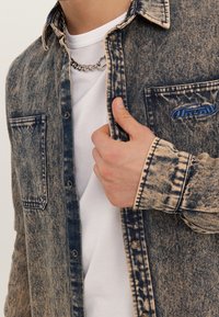 Koszula jeansowa w wyblakłym niebieskim kolorze z kontrastującym beżowym przeszyciem, zapinana na zatrzaski, z dwoma kieszeniami na piersi i małą niebieską naszywką z logo.